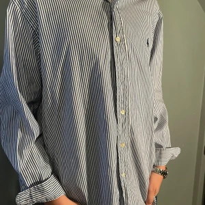 Randig skjorta från Ralph Lauren - Klassisk blåvit randig skjorta från Ralph Lauren i modellen Classic Fit. Modellen är 177-178 cm så den kommer att passa både M och L beroende på hur man vill ha pass formen. Skjortan har långa ärmar, knappar framtill och broderad logga på bröstet. Tillverkad i mjuk bomull och passar perfekt för en clean och stilren look.