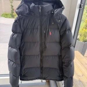 Svart dunjacka från Polo Ralph Lauren - Säljer en svart dunjacka från Polo Ralph Lauren med huva och dragkedja. Jackan har en klassisk pufferstil, diskret logga i rött på bröstet och flera praktiska fickor med dragkedja. Perfekt för kalla dagar och riktigt snygg till streetwear.