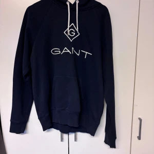 Mörkblå hoodie från GANT XL - Säljer en mörkblå hoodie från GANT i storlek XL. Tröjan har vit broderad logga på bröstet, huva med snörning och en stor magficka. Perfekt för dig som gillar klassisk och stilren streetwear.