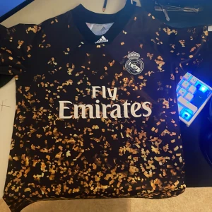 Real Madrid Adidas svart/guld tröja M - Snygg Real Madrid fotbollströja från Adidas i svart med guldmönster och vitt tryck. Tröjan har korta ärmar, klubbmärke på bröstet och Fly Emirates-logga. Tillverkad i lätt och ventilerande Climalite-material.
