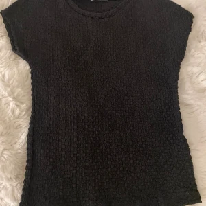 Svart strukturerad topp från Zara - Säljer en svart topp från Zara i storlek S. Toppen har en cool strukturerad yta och rund hals. Den är kortärmad och har en slim passform som sitter snyggt på kroppen. Perfekt för dig som gillar stilrena och trendiga plagg.