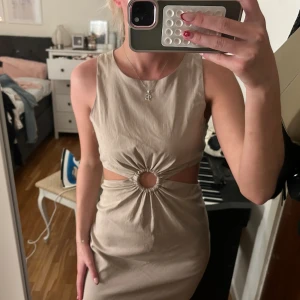 Beige cut out klänning från Zara - Säljer en beige kort klänning från Zara med snygga cut outs i midjan och en rund detalj framtill. Klänningen är ärmlös och har en figurnära passform. Perfekt för dig som vill sticka ut med en trendig och stilren look.