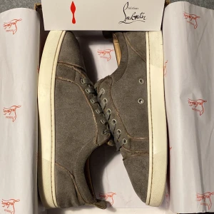 Grå sneakers från Christian Louboutin - Snygga grå sneakers från Christian Louboutin med klassisk röd sula. Skorna har rund tå, snörning och är tillverkade i canvas med detaljer i skinn. Perfekta för dig som vill ha en stilren och exklusiv look med en ikonisk touch.