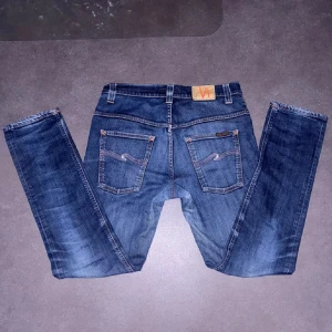 Blå jeans från Nudie Jeans - Säljer ett par klassiska blå jeans från Nudie Jeans med snygga kontrastsömmar och ikonisk broderad bakficka. Jeansen har normal passform och raka ben, tillverkade i slitstarkt denimtyg av bomull. Perfekta för dig som gillar en tidlös och avslappnad look. 33/32