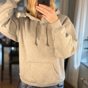 Grå hoodie från Bik Bok med huva - Mysig grå hoodie från Bik Bok i storlek XS. Tröjan har huva med snörning, magficka och ribbade muddar vid ärmslut och nederkant. Perfekt för chill dagar eller när du vill ha en enkel och stilren look.