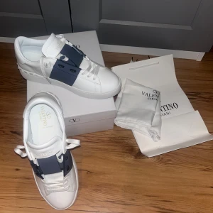 Valentino Garavani Open sneakers vit/blå - Snygga Valentino Garavani Open sneakers i vitt skinn med bred blå rem över vristen och coola nitar på sulan. Klassisk rund tå, vita snören och diskret guldlogga på plösen. Perfekta för dig som vill ha en stilren men ändå unik look.