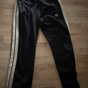 Nya palm angels byxor  - 100% authentic Svarta palm angels trackpants med vita sidoränder och svart linje, dragkedja vid benslut och broderad bokstav på höften. Snygg sportig look med elastisk midja och raka ben. Perfekta för chill eller streetwear.