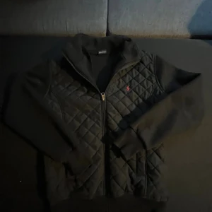 Svart quiltad jacka Polo Ralph Lauren - Svart quiltad jacka från Polo Ralph Lauren med dragkedja framtill och klassisk röd broderad logga på bröstet. Jackan har långärmade ärmar i mjukt material och en hög krage. Perfekt för dig som gillar stilren och sportig look.