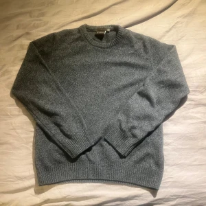 Grå stickad tröja från Oscar Jacobson - Mysig grå stickad tröja från Oscar Jacobson i klassisk modell. Tröjan har rund halsringning, ribbade muddar och är tillverkad i 80% ull och 20% polyamid. Perfekt för lager-på-lager under kyliga dagar.