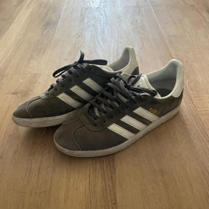 Adidas Gazelle sneakers - Säljer ett par klassiska Adidas Gazelle sneakers i mörkgrå mocka med vita detaljer och de ikoniska tre ränderna på sidorna. Skorna har snörning, platt sula och rund tå. Perfekta för dig som gillar retrostil och streetwear.
