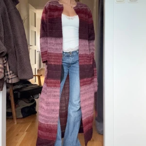 Randig lång cardigan i vinrött - Säljer en supermysig lång cardigan med breda ränder i vinrött, mörkrött och rosa toner. Jackan är stickad, har lång ärm och ett bälte i midjan & har även fickor🩷❤️Perfekt att slänga över en outfit som funkar året runt. Materialet är fluffigt och mjukt, och jackan har en loose passform. Kommer ifrån Julie Fagerholt - Heartmade (dansk fashion designer)