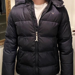 Mörkblå pufferjacka från Gant - Säljer en mörkblå pufferjacka från Gant med huva och dragkedja framtill. Jackan har en klassisk quiltad look, Gant-logga på bröstet och snygg röd detalj på insidan. Perfekt för kalla dagar och riktigt skön att bära.