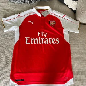 Arsenal Puma fotbollströja röd/vit - Arsenal fotbollströja från Puma i rött och vitt med klubbmärke på bröstet och Fly Emirates-tryck. Klassisk krage med knapp, guldiga detaljer och korta ärmar. Tillverkad i lätt och ventilerande polyester, perfekt för match eller träning.