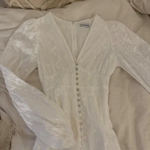 Vit långklänning - Superfin vit långklänning med broderade blommor, v-ringning och långa puffärmar. Klänningen har en rad snygga knappar framtill och är gjord i ett lätt bomullstyg. Använd en gång på student, från Adoore