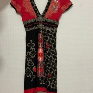 Mönstrad klänning från Desigual - Färgstark kortklänning från Desigual med svart, rött och beige blommönster och grafiska detaljer. Djup v-ringning, korta ärmar och kontrasterande mönster gör den unik. Perfekt för dig som gillar statement-plagg och lekfull design.