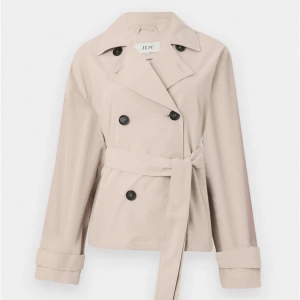 Beige trenchcoat från JDY med bälte - Snygg beige trenchcoat från JDY med klassisk krage, dubbelknäppning och bälte i midjan. Jackan har långa ärmar med knäppdetalj vid mudden och en stilren, kortare passform. Perfekt för dig som gillar minimalistisk och clean stil.