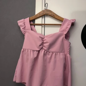 Rosa volangtopp från SHEIN - Supersöt rosa topp från SHEIN med breda axelband och volangdetaljer. Toppen har rynk framtill och smockad rygg för extra komfort. Den är ärmlös och har en luftig, flowy passform som känns perfekt för varmare dagar.