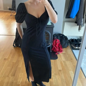 Svart långklänning från Gina Tricot - Supersnygg svart långklänning från Gina Tricot med puffärmar, v-ringning och knyt framtill. Klänningen har en slits på sidan som ger en extra edge och är figurnära i modellen. Perfekt för dig som gillar trendiga och feminina detaljer.