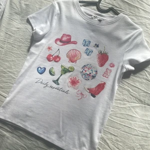 Vit t-shirt med färgglada tryck från FB Sister - Vit t-shirt från FB Sister i storlek XS med färgglada tryck på framsidan, bland annat cowboyhatt, körsbär, discokula och boots. Modellen har rund hals och korta ärmar. Materialet är mjuk bomull och passformen är normal. Perfekt för dig som gillar roliga detaljer.