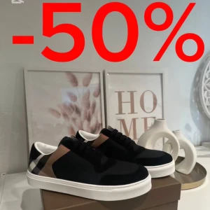 Burberry sneakers svart/vit - Snygga Burberry sneakers med klassiskt rutigt mönster i svart, beige och vitt på hälen. Ovandel i svart textil och mocka, vita sulor och svarta skosnören. Perfekta för dig som vill ha en stilren och lyxig look med ikoniska Burberry-detaljer.