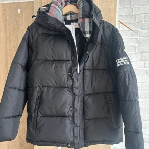 Svart pufferjacka från Burberry - Svart pufferjacka från Burberry med huva och dragkedja framtill. Jackan har Burberry-logga på ärmen och klassiskt rutigt foder i grått, vitt och rött. Två fickor med dragkedja och quiltad design ger en modern vibe. Perfekt för kalla dagar.