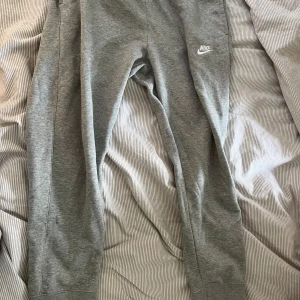 Grå mjukisbyxor från Nike - Oanvända! Snygga grå mjukisbyxor från Nike med broderad vit logga på låret. Byxorna har en klassisk passform med resår i midjan och muddar vid bensluten. Perfekta för chill eller träning, tillverkade i mjukt bomullsmaterial för extra komfort.