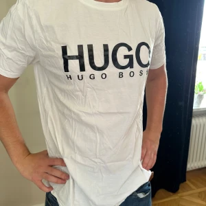 Vit Hugo Boss t-shirt med tryck - Vit t-shirt från Hugo Boss med stor svart logotyp på bröstet. Klassisk rund hals och korta ärmar. Tillverkad i mjuk bomull som känns skön mot huden. Perfekt för dig som gillar stilrena och enkla plagg med tydlig branding.
