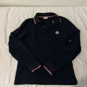 Mörkblå långärmad pikétröja Moncler - Snygg mörkblå långärmad pikétröja från Moncler med klassisk krage och knäppning framtill. Tröjan har Moncler-logga på bröstet och detaljer i rött och vitt på krage och ärmslut. Tillverkad i bomull för en skön känsla.