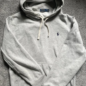 Grå hoodie från Polo Ralph Lauren - Snygg grå hoodie från Polo Ralph Lauren med klassisk blå logobrodyr på bröstet. Tröjan har huva med snörning, ribbade muddar och en mjuk insida. Perfekt för chill dagar och enkel att matcha med jeans eller joggers.