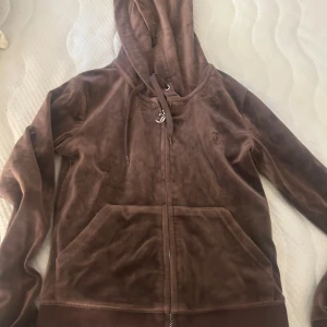 Brun velour hoodie från Juicy Couture - Mysig brun hoodie från Juicy Couture i mjuk velour med dragkedja framtill, huva med snörning och känguruficka. Broderad JC-logga på bröstet. Perfekt för chill dagar eller när du vill ha en avslappnad men snygg look.