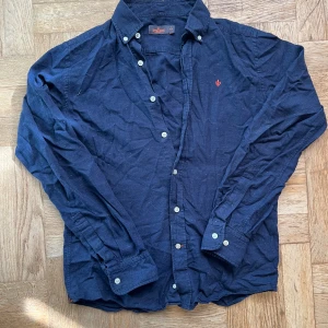 Mörkblå skjorta från Morris - Säljer en mörkblå skjorta från Morris i bomull med små färgstänk och klassisk krage. Skjortan har knappar hela vägen, lång ärm och en broderad orange logga på bröstet. Perfekt för dig som gillar stilrena detaljer och snygg passform.