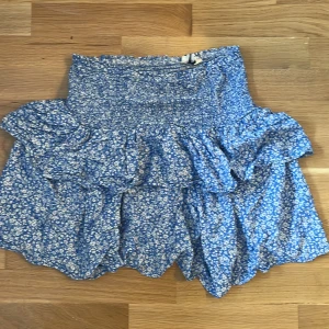 Blå blommig volangkjol från Kappahl - Supersöt blå kortkjol från Kappahl med vitt blommönster och volangdetaljer. Kjolen har smock i midjan för en bekväm passform och är gjord i ett lätt och luftigt material. Perfekt för dig som gillar en feminin och somrig stil! Helt okej skick, använd få gånger 