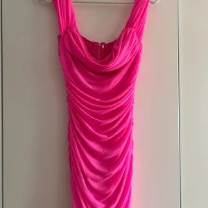 Rosa draperad kortklänning - Supersnygg rosa kortklänning med draperad effekt och breda axelband. Klänningen har en snygg cowlneck-ringning och dragkedja bak. Perfekt för dig som vill sticka ut med en färgstark look.