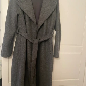 Grå lång kappa med bälte - Snygg och stilren grå kappa med bälte i midjan och breda slag. Lång modell med raka ärmar och två fickor framtill. Perfekt för kyligare dagar och ger en klassisk look.