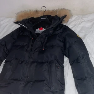 Svart pufferjacka från Zavetti Canada - Säljer en svart pufferjacka från Zavetti Canada med fet pälskant på huvan och snyggt broderat märke på ärmen. Jackan har dragkedja, knappar och flera fickor framtill. Perfekt för kalla dagar och riktigt skön att bära.