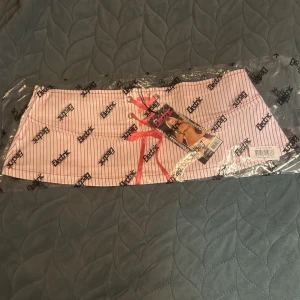 Randig minikjol från Electric Lingerie - Supersöt kortkjol från Electric Lingerie i vitt med tunna röda ränder. Kjolen har snörning framtill med rosa band och små rosetter bak. Tillverkad i mjuk bomull med lite stretch för perfekt passform. Perfekt för dig som vill sticka ut med en lekfull och trendig look.