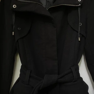 Svart kappa med bälte och dragkedja - Stilren svart kappa med bälte i midjan och dragkedja framtill. Jackan har stora fickor, långa ärmar och snygga detaljer vid axlarna. Perfekt för dig som vill ha en klassisk look med modern touch.