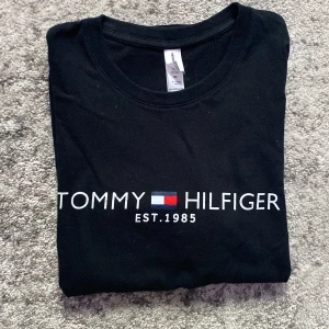 Svart t-shirt Tommy Hilfiger print - Svart t-shirt i bomull med klassiskt Tommy Hilfiger-tryck och logga framtill. Rund halsringning och normal passform. Perfekt för dig som gillar stilrena och ikoniska plagg med tydlig branding.
