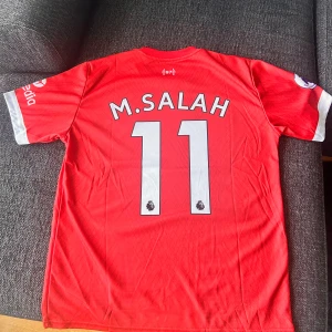 Liverpool  - Liverpool FCs officiella matchtröja. Salah och nummer 11 på ryggen. Röd med vita detaljer, kortärmad och tillverkad i lätt Dri-FIT-material. Nike-logga, LFC-märke och Premier League-patch på ärmen. Perfekt för alla fans! Large men sitter som medium