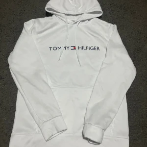 Vit hoodie från Tommy Hilfiger M  - Snygg vit hoodie från Tommy Hilfiger i storlek M. Tröjan har klassisk huva med snörning, stor magficka och tryckt logga framtill. Perfekt för dig som gillar stilrena och bekväma plagg. Materialet är mjukt och känns skönt mot huden.
