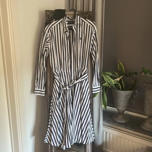 Randig skjortklänning från Polo Ralph Lauren - Snygg skjortklänning från Polo Ralph Lauren i vitt och marinblått med vertikala ränder. Klänningen har lång ärm, knappar framtill, klassisk krage och avtagbart bälte i midjan. Tillverkad i bomull och har broderad logga på bröstet. Perfekt för en fräsch och stilren look.