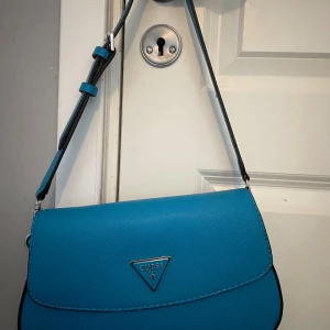 Blå axelväska från Guess - Snygg blå axelväska från Guess med justerbart axelband och klassisk trekantslogga i silver framtill. Väskan är tillverkad i strukturerad syntet och har en stilren, modern design som passar perfekt till din outfit.