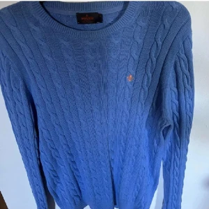 Blå kabelstickad tröja från Morris - Kabelstickad blå tröja från Morris i 100% bomull. Klassisk rund halsringning, ribbade muddar och diskret broderad logga på bröstet. Perfekt för dig som gillar stilrena och tidlösa plagg med snygg struktur.