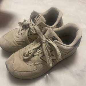 Beige New Balance 574 sneakers - Säljer ett par klassiska New Balance 574 sneakers i beige med svarta detaljer. Skorna har en ikonisk N-logga på sidan, snörning och en mix av mocka och textilmaterial. Perfekta för dig som gillar retrostil och vill ha bekväma sneakers till vardags. Använd några veckor tills de kändes lite för små. 