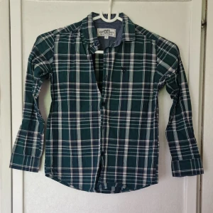 Grönrutig skjorta från Hampton Republic - Snygg grön och vit rutig skjorta från Hampton Republic i storlek 128. Skjortan har klassisk krage, långa ärmar och knäppning framtill. Perfekt för en chill och stilren look. Tillverkad i mjuk bomull som känns skön mot huden.