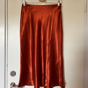 Rostfärgad/oreange satinkjol  - Snygg midikjol i skimrande rostfärgad/orange färg. Kjolen faller mjukt med en lätt utsvängd siluett. Dragkedja i sidan, midjemått 70 cm, passar s/m. Köpt second hand. Säljer en liknande i brunt också! 