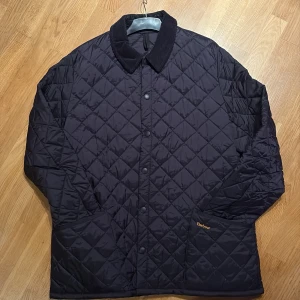 Svart quiltad jacka från Barbour XXL - Quiltad svart jacka från Barbour i storlek XXL. Jackan har klassisk krage i manchester, tryckknappar framtill och Barbour-logga broderad på fickan. Perfekt för dig som gillar stilren och tidlös design.