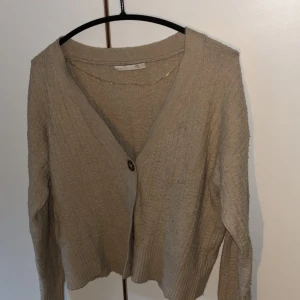 Beige kofta från 157 med knapp - Beige stickad kofta från 157 med v-ringning och en stor knapp framtill. Koftan har långärmad design, ribbade muddar och en avslappnad passform. Perfekt för lager-på-lager och chill dagar.