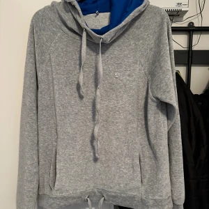 Grå hoodie i velour med blå huva strl: M - Grå hoodie i velour med mjukt material och blå insida i huvan. Tröjan har snörning både vid huvan och nederkanten, samt en liten logga på bröstet. Långärmad och med praktiska fickor på sidorna. Perfekt för chill dagar eller när du vill vara extra comfy. Loggan är något sprucket. Förekommer noppor.