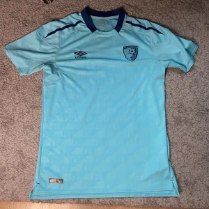 Bournemouth fotbollströja - Snygg ljusblå AFC Bournemouth fotbollströja från Umbro med klubbmärke på bröstet och mörkblå detaljer vid kragen och axlarna. Tillverkad i lätt och ventilerande polyester, perfekt för match eller träning. Officiell licensierad produkt.